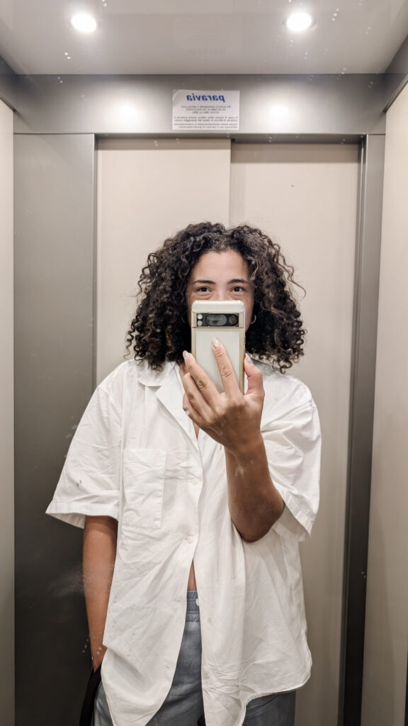 Fashion-forward elevator selfie of a young woman in Maiori, Italy, wearing an elevated casual outfit with a boyfriend shirt and satin trousers, redefining postpartum style on the Amalfi Coast.