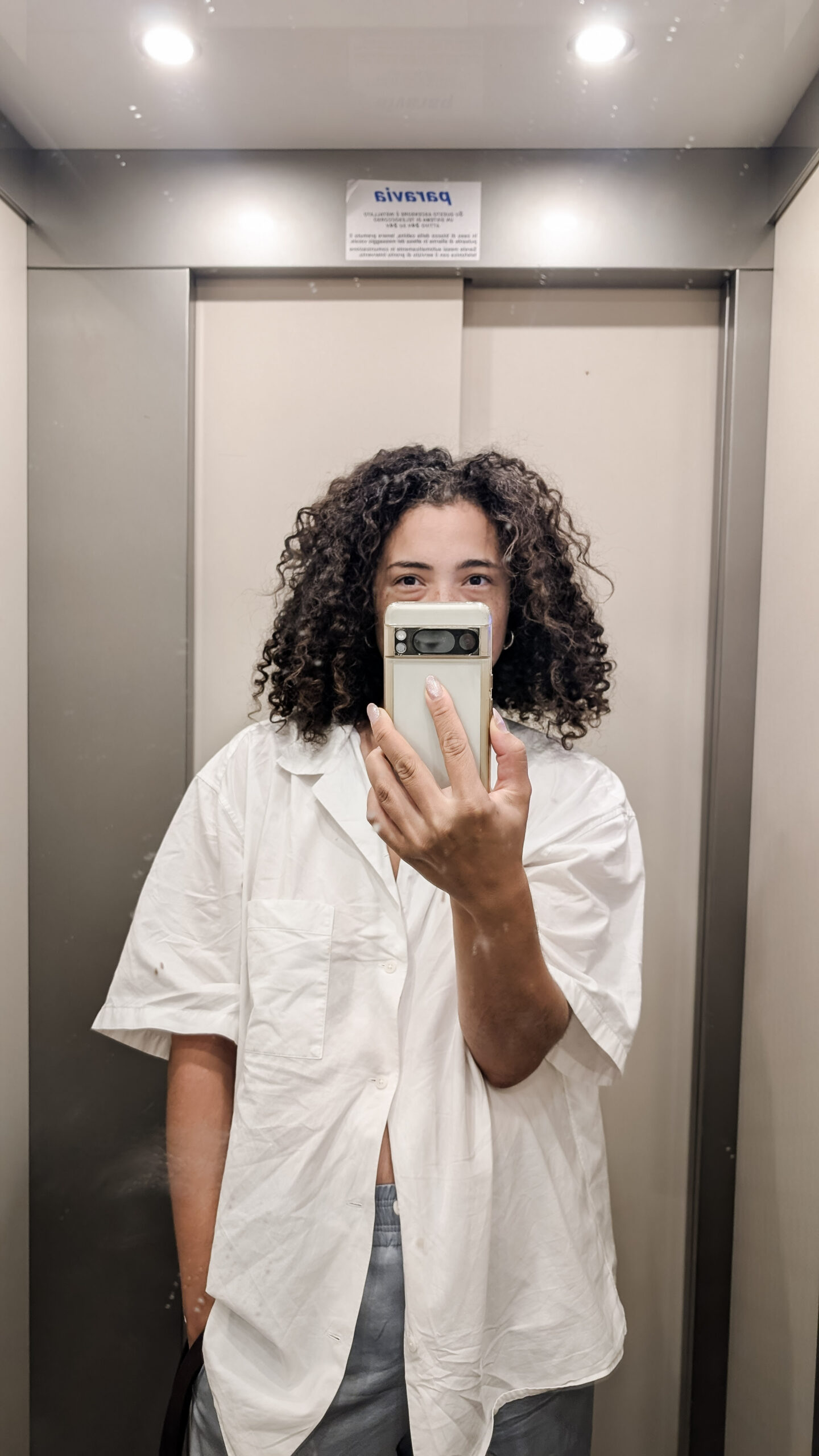 Fashion-forward elevator selfie of a young woman in Maiori, Italy, wearing an elevated casual outfit with a boyfriend shirt and satin trousers, redefining postpartum style on the Amalfi Coast.